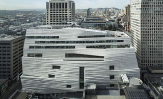 Exterior view of the off-white coloured, irregular shaped San Francisco Museum of Modern Art (SFMoMA) building during the day. There are multiple buildings surrounding the museum