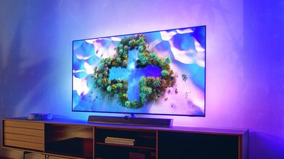 Philips OLED+936 sitting on wooden TV bench, with Ambilight light spreading onto the wall behind it