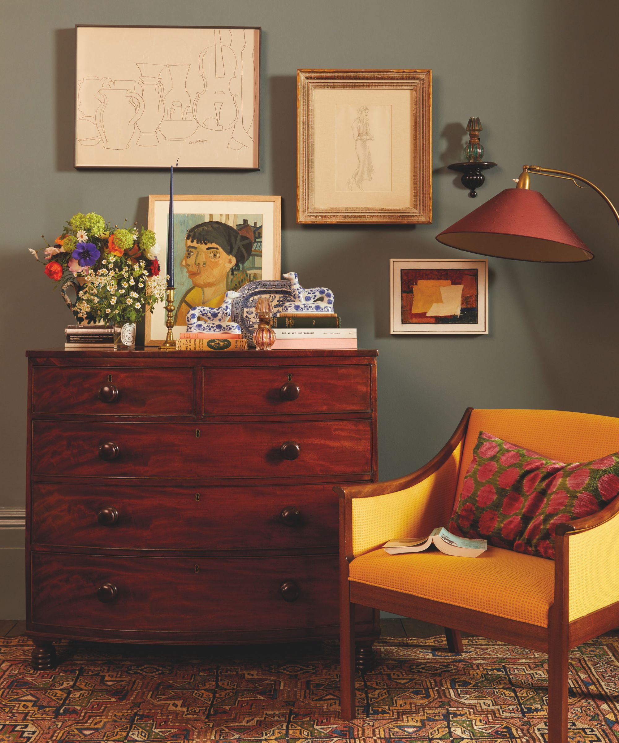 A vintage mahogany chest of drawers against a dark moody green wall, decorated with framed sketches, colorful flowers, and blue-and-white ceramic figurines