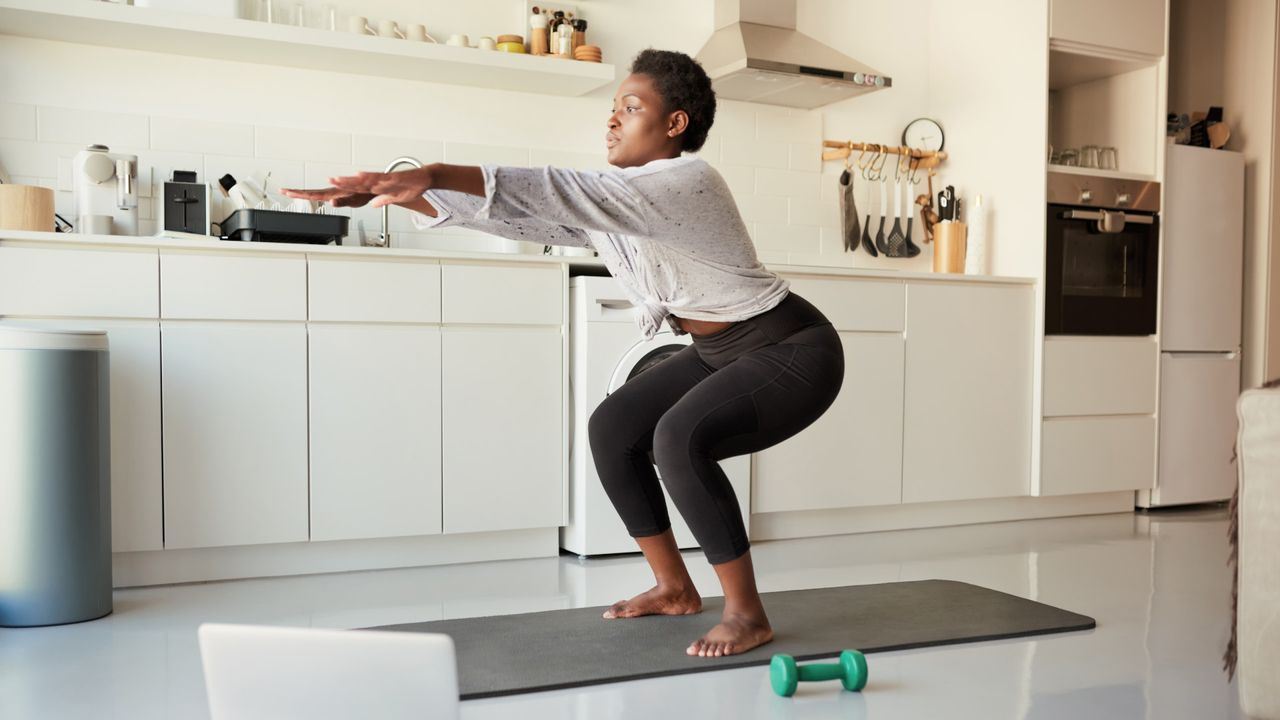 A woman performing a squat following a Pilates workout on her laptop