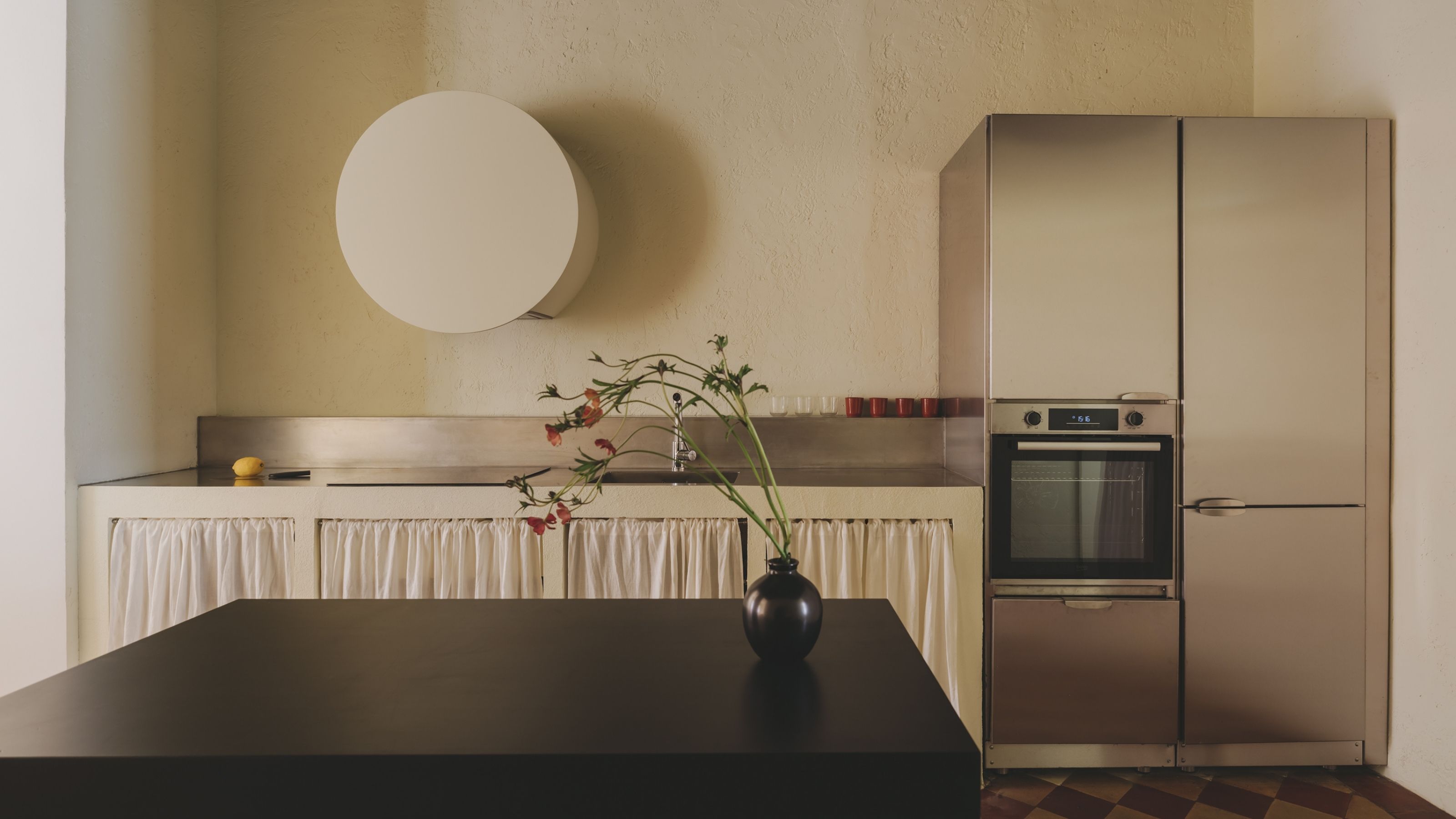 A butter yellow-tinted kitchen with a black wood kitchen island topped by a ceramic vase, matte chrome kitchen countertops, white curtains to conceal shelving, and a round, white lighting bulb.