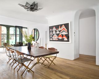 A dining room with a six seater table, wooden chairs, a vase with plants in, a large wall painting and wooden floors.