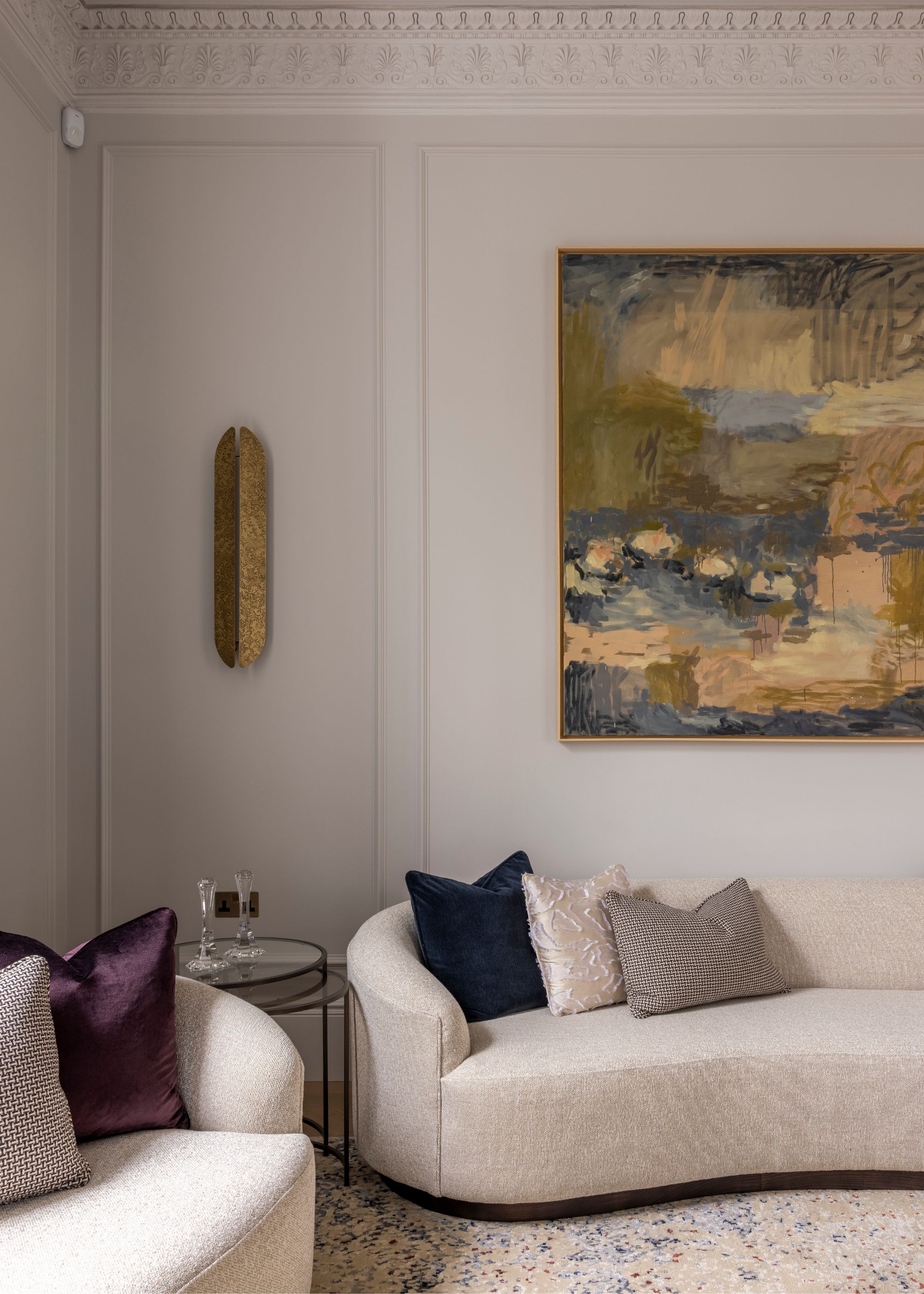 A modern living room with light gray panelled walls, a large piece of abstract art on the wall in a green color palette, and a curved, cream sofa.