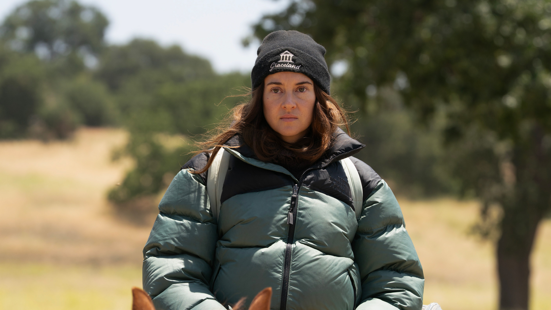 A woman (Shailene Woodley as Annie Clay) wears a black "Graceland" beanie and a puffy green jacket while riding atop a horse outdoors, in 'Paradise' season 2.