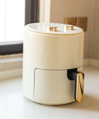 A white circular air fryer with a gold handle on the drawer, standing on a white windowsill with a white wall and window behind it