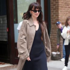 Dakota Johnson steps outside in New York wearing cream heels with a navy dress and a dark beige trench coat.