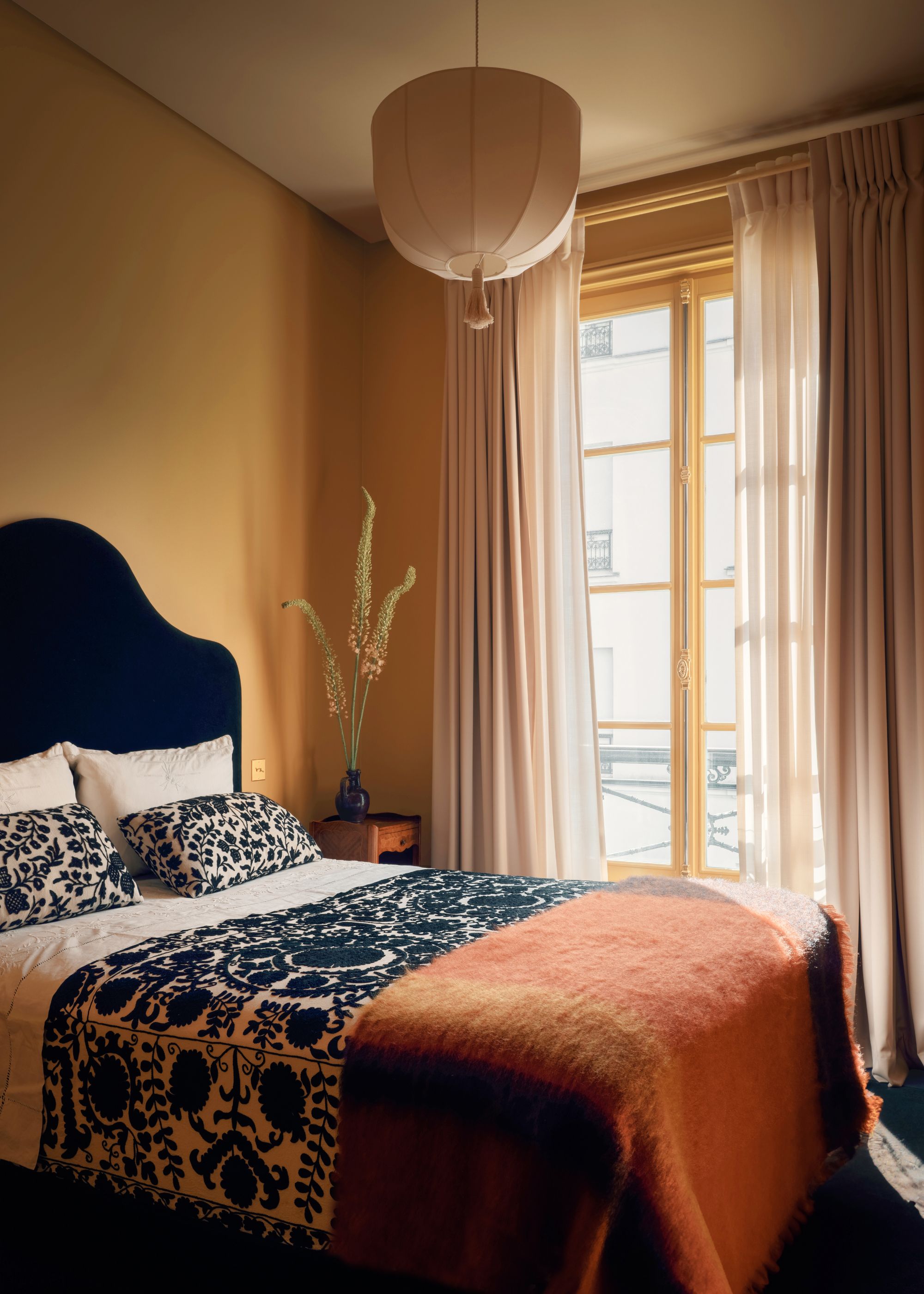 A bedroom with dark yellow walls, a beige ceiling, and French doors leading onto a balcony. Sheer beige curtains over the doors, and a bed with a black upholstered headboard, cream and black patterned cushions, a throw, and an orange-red blanket.