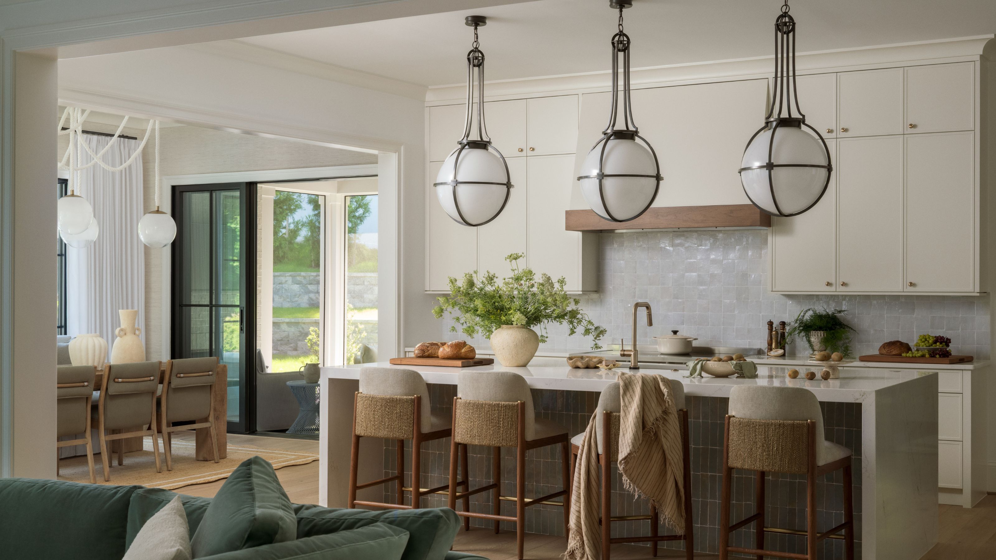 A bright kitchen featuring a large waterfall island with woven-back stools, three oversized globe pendant lights with black metal frames, and a white tiled backsplash.