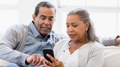 An older couple looks at a smartphone together while sitting on their sofa.