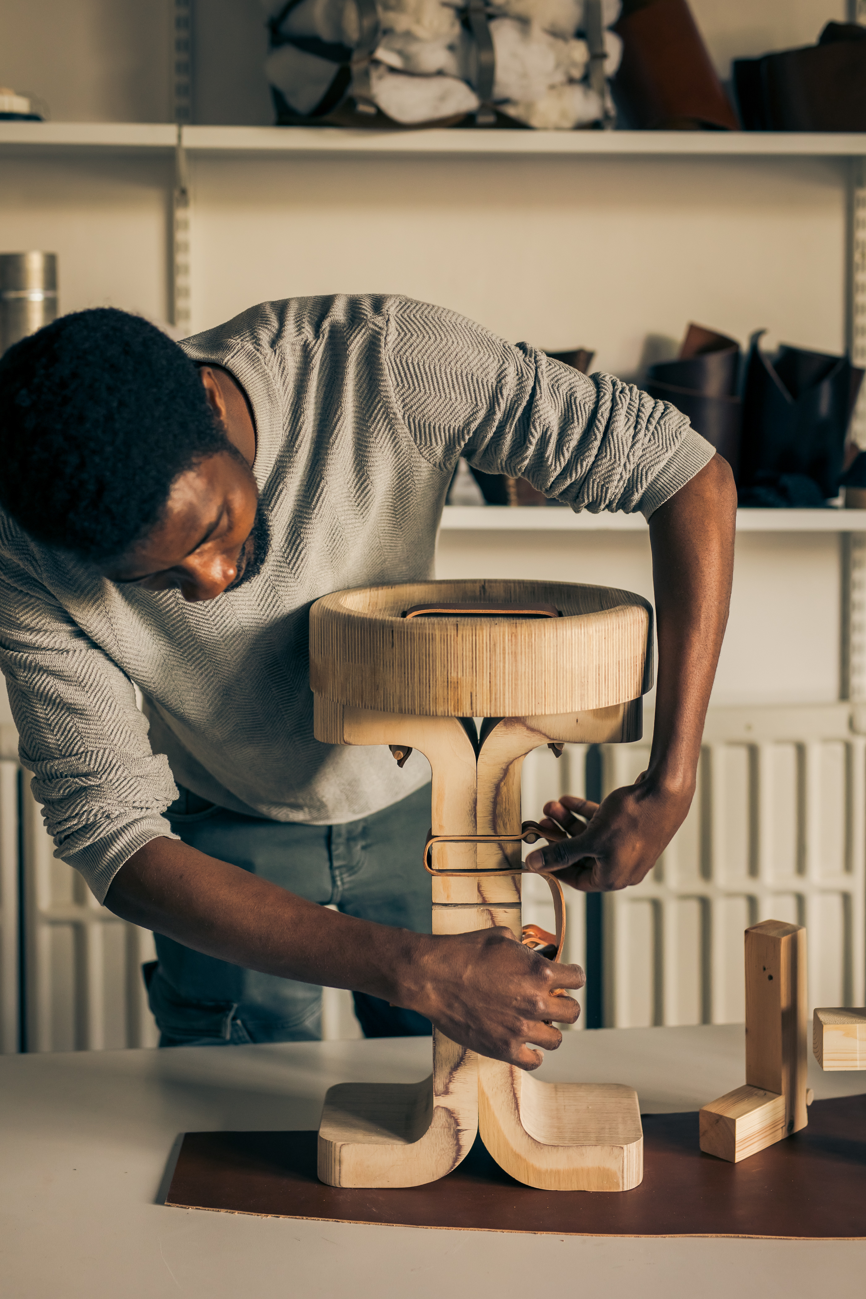 A young woman wearing a grey long-sleeved shirt and dark blue trousers handles a wooden stool that&#039;s being crafted manually in a white, industrial studio space.
