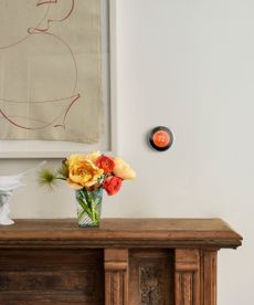 A small round thermostat with an orange screen on a cream painted wall beside a large contemporary artwork. Above a dark wood sideboard with a vase of yellow and red flowers on it. 