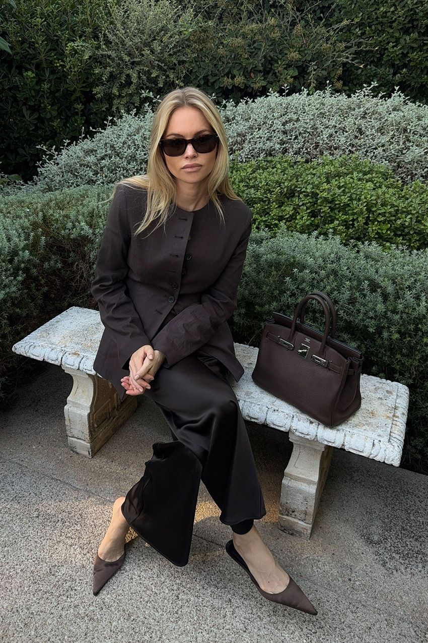 Fall shoe trends in Paris are shown in a photo of a woman sitting at a park wearing the satin shoe trend with a brown blazer, black satin trouser, and brown satin kitten heels