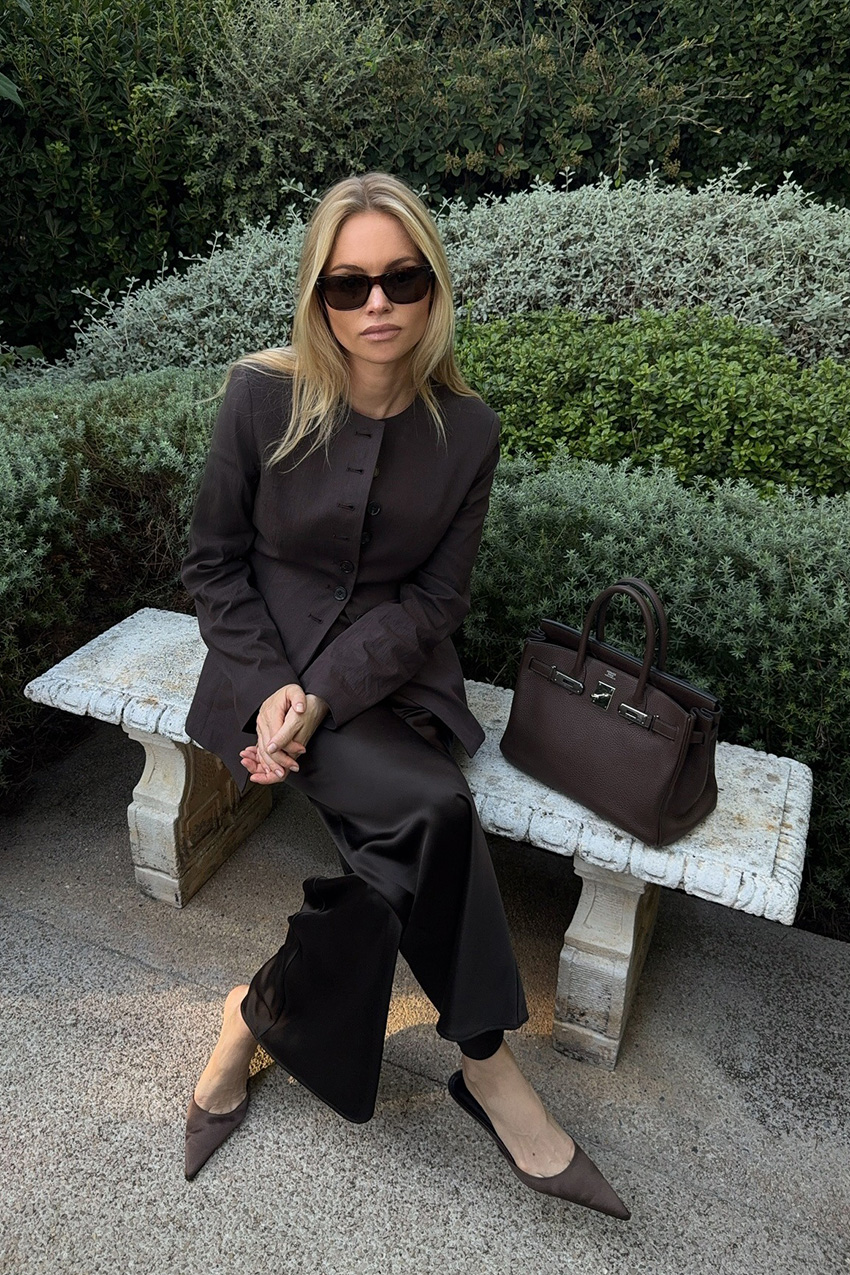 Fall shoe trends in Paris are shown in a photo of a woman sitting at a park wearing the satin shoe trend with a brown blazer, black satin trouser, and brown satin kitten heels
