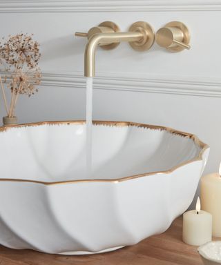 A white scalloped countertop basin with gold trim sits on a wooden surface. A brushed gold wall-mounted tap dispenses water. Nearby, lit candles and dried flowers add warmth.