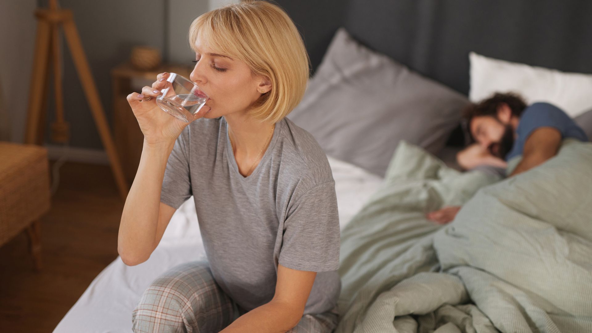 A woman sat up in bed drinking a glass of water next to a man who's asleep