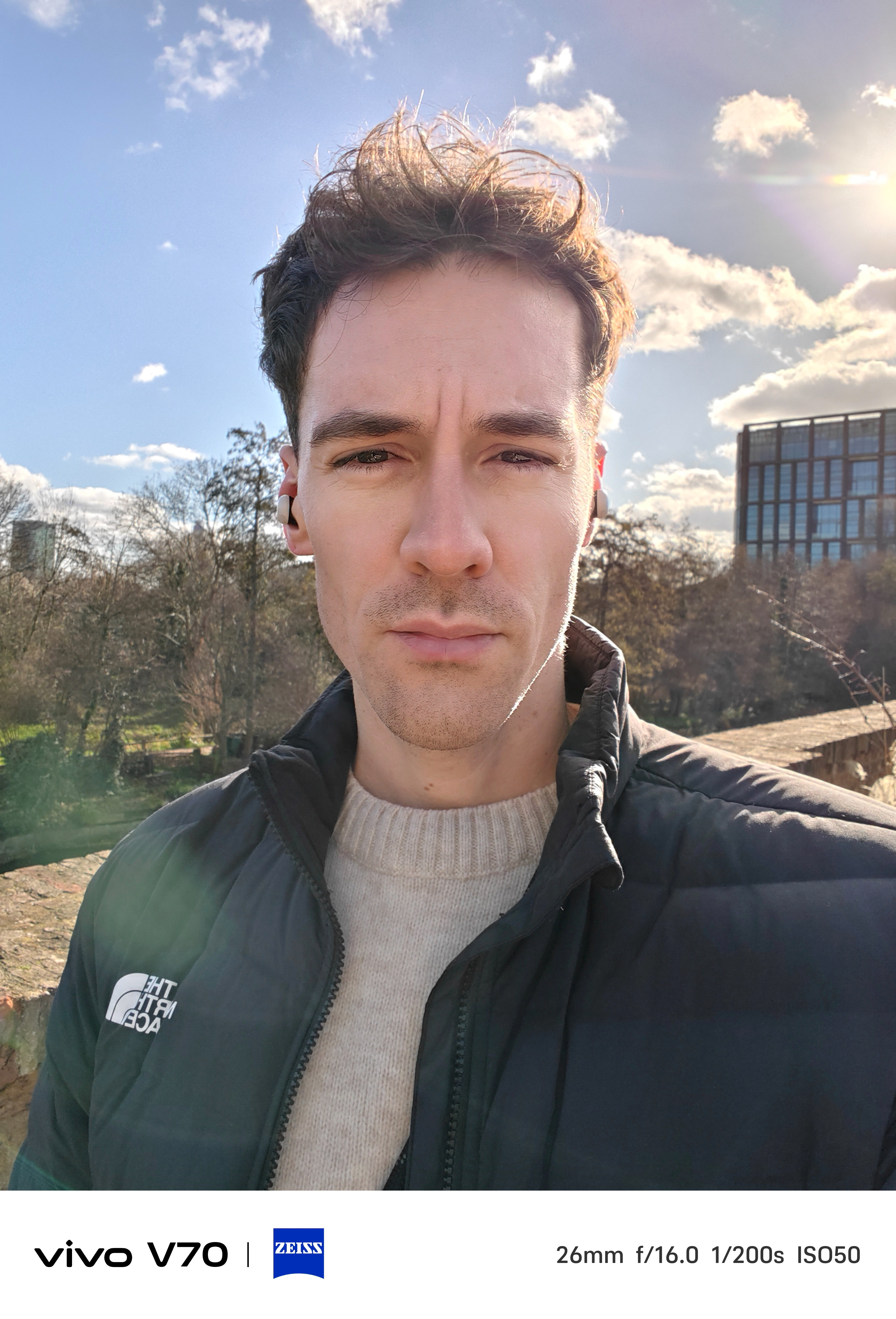 Outdoor selfie of a man in bright sunlight with blue sky and modern building behind him.