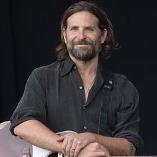 Bradley Cooper poses with a guitar on his lap and microphone on a stand.