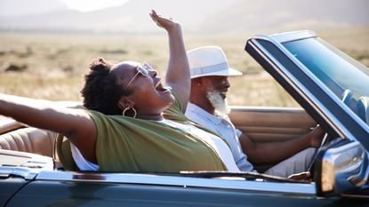 A middle-aged couple in a convertible look like they're having a great time driving down the road.
