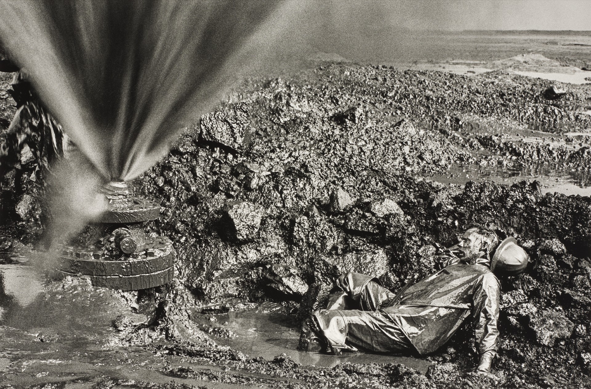 A black-and-white photograph of a figure in a reflective protective suit and helmet lying on oil-soaked rocky ground, with a pressurised jet of gas erupting from a wellhead to the left and a flat, dark horizon stretching behind.