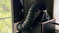 A black flexible exhaust hose connects a computer tower to a window vent, with a tall black tower fan standing vertically beside the window frame.