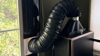 A black flexible exhaust hose connects a computer tower to a window vent, with a tall black tower fan standing vertically beside the window frame.