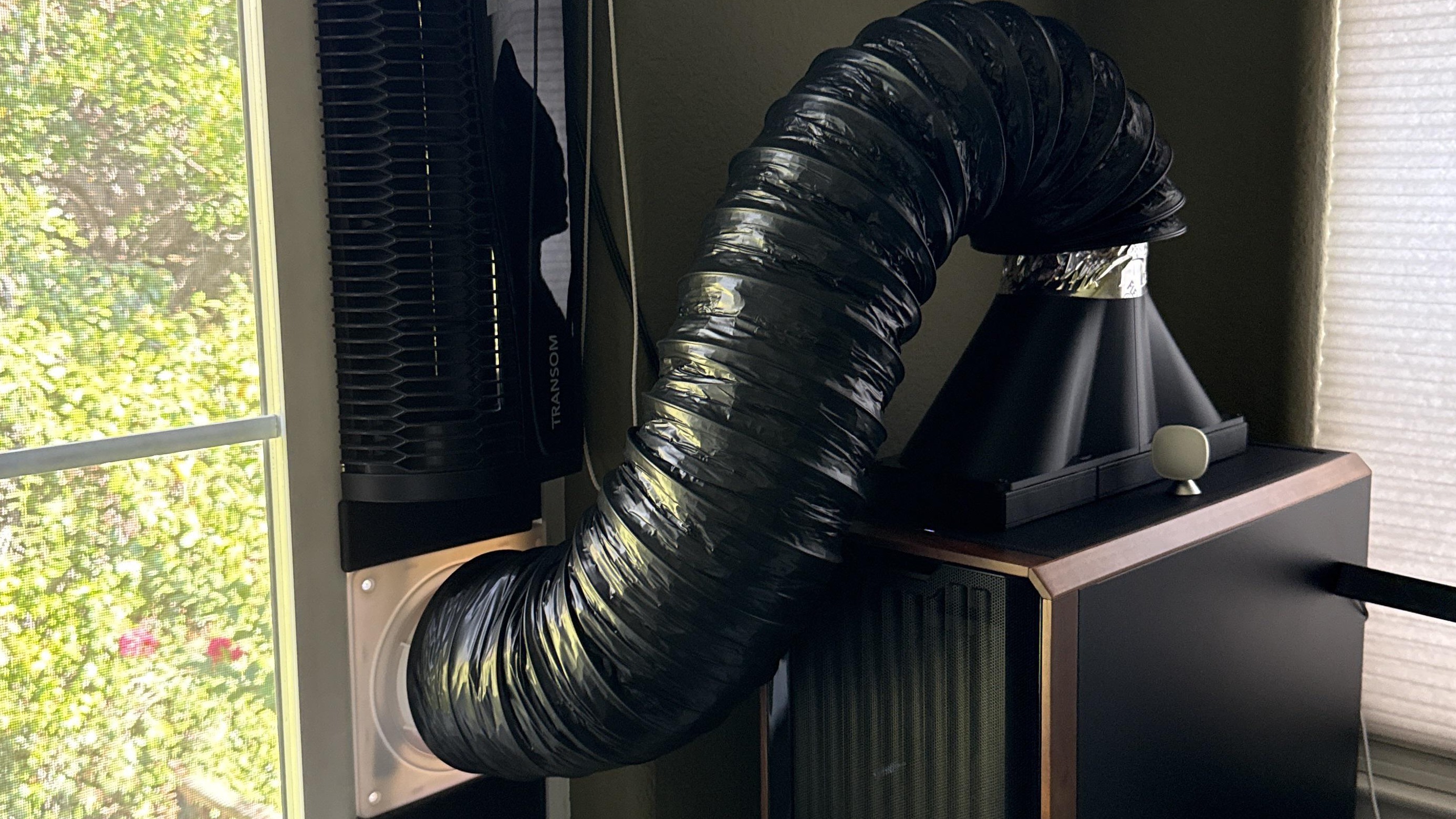 A black flexible exhaust hose connects a computer tower to a window vent, with a tall black tower fan standing vertically beside the window frame.