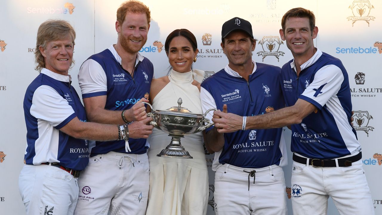 Prince Harry and Meghan Markle posing with polo players holding a trophy in front of a white background