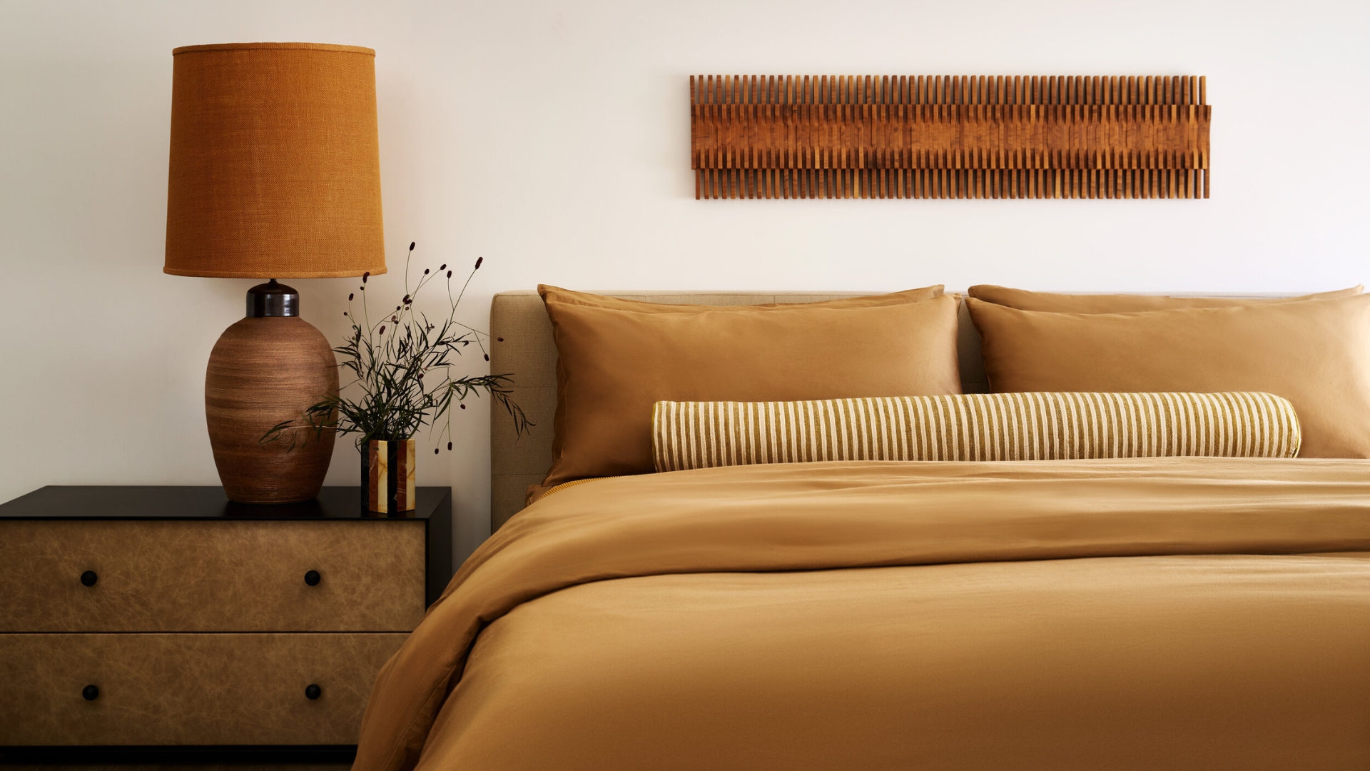 A white bedroom with an ochre bedscape, a striped bolster pillow, a wood side table with a tall table lamp, a vase of stems, and a textured wall hanging