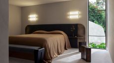 A bedroom with a pair of wall sconces above the oversized headboard on either side, lighting up the brown bedscape below. Also pictured, a bench at the foot of the bed and a pair of wood blocks beside