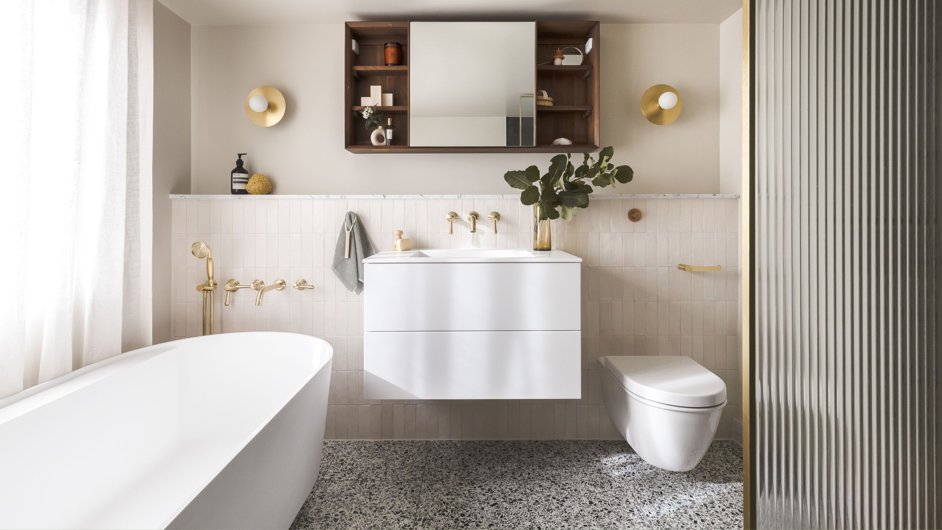 A bathroom that has been zoned up. You can see a view of the bathtub, the bathroom vanity and sink as well as the toilet and a split area for the shower. Above the bathroom sink is a large mirrored cabinet with open shelving on either side of it. 