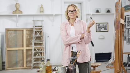 A confident-looking older woman stands in her art studio holding a paintbrush.