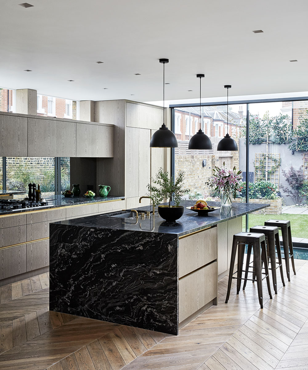 A black marble kitchen island with metal stools in a luxurious kitchen extension with herringbone wood flooring.