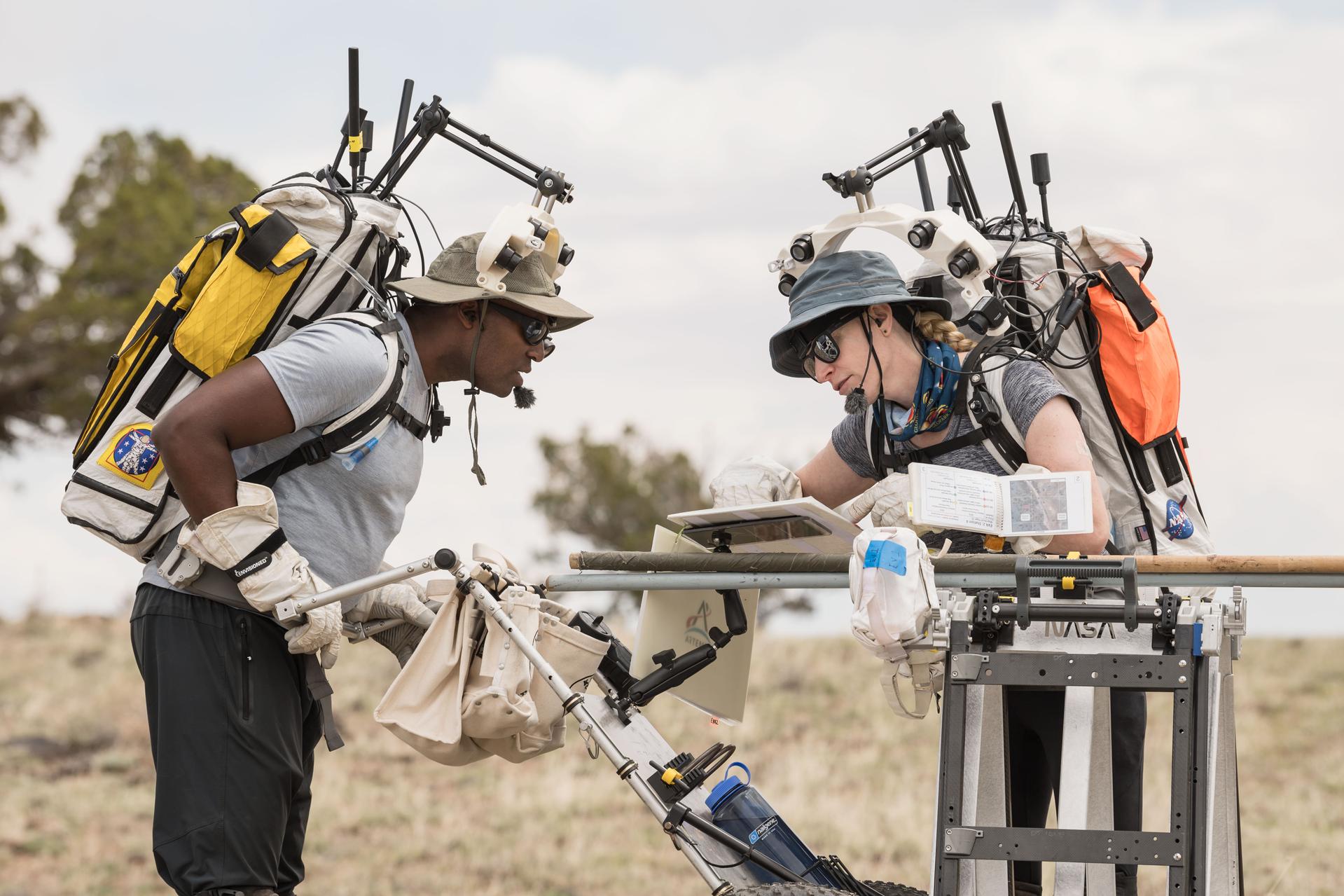 two astronauts leaning over a table in a field, while wearing backpacks with antennas on top