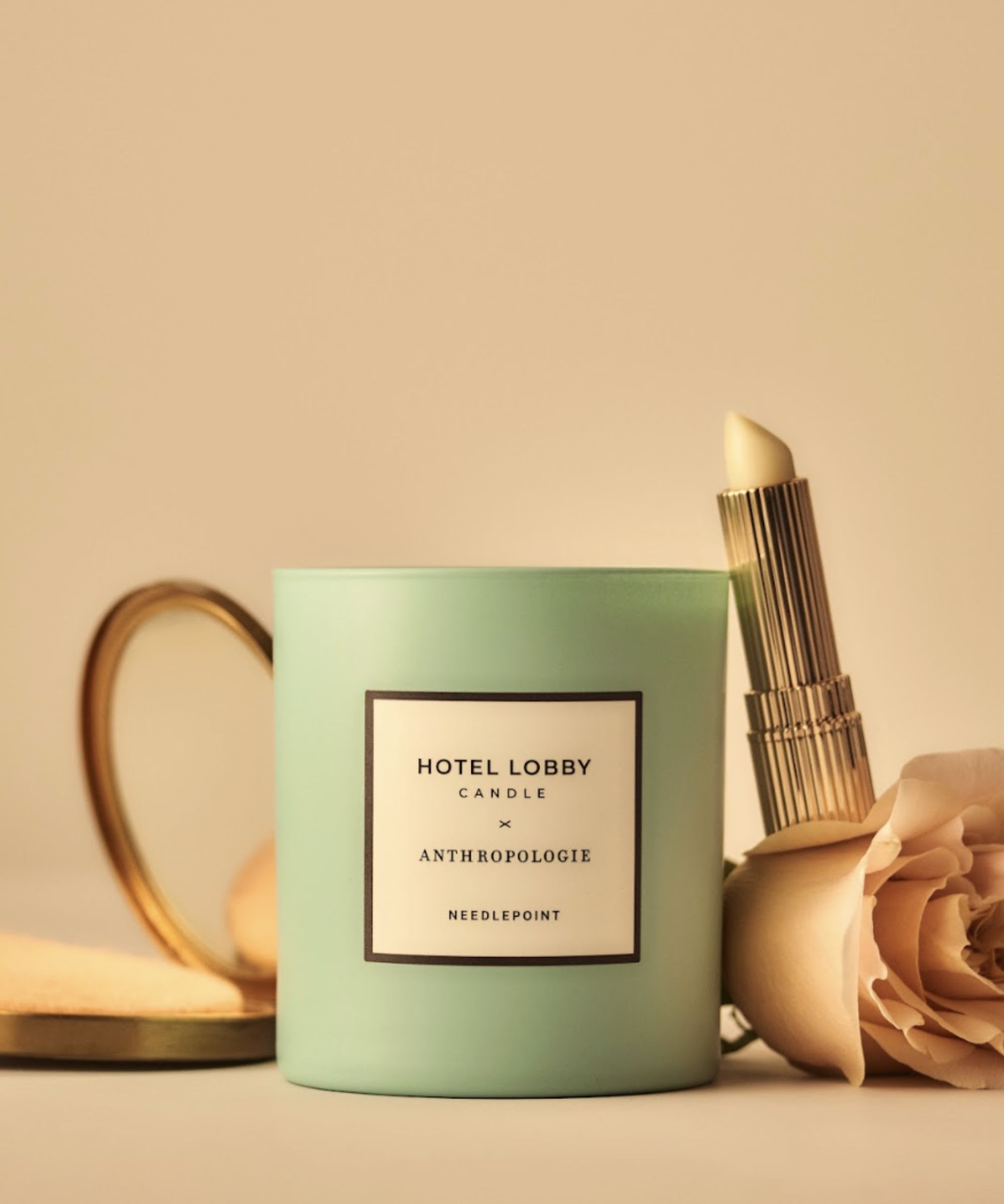 A pale green Hotel Lobby candle against a soft peach background, pictured with a pale pink rose and gold lipstick.