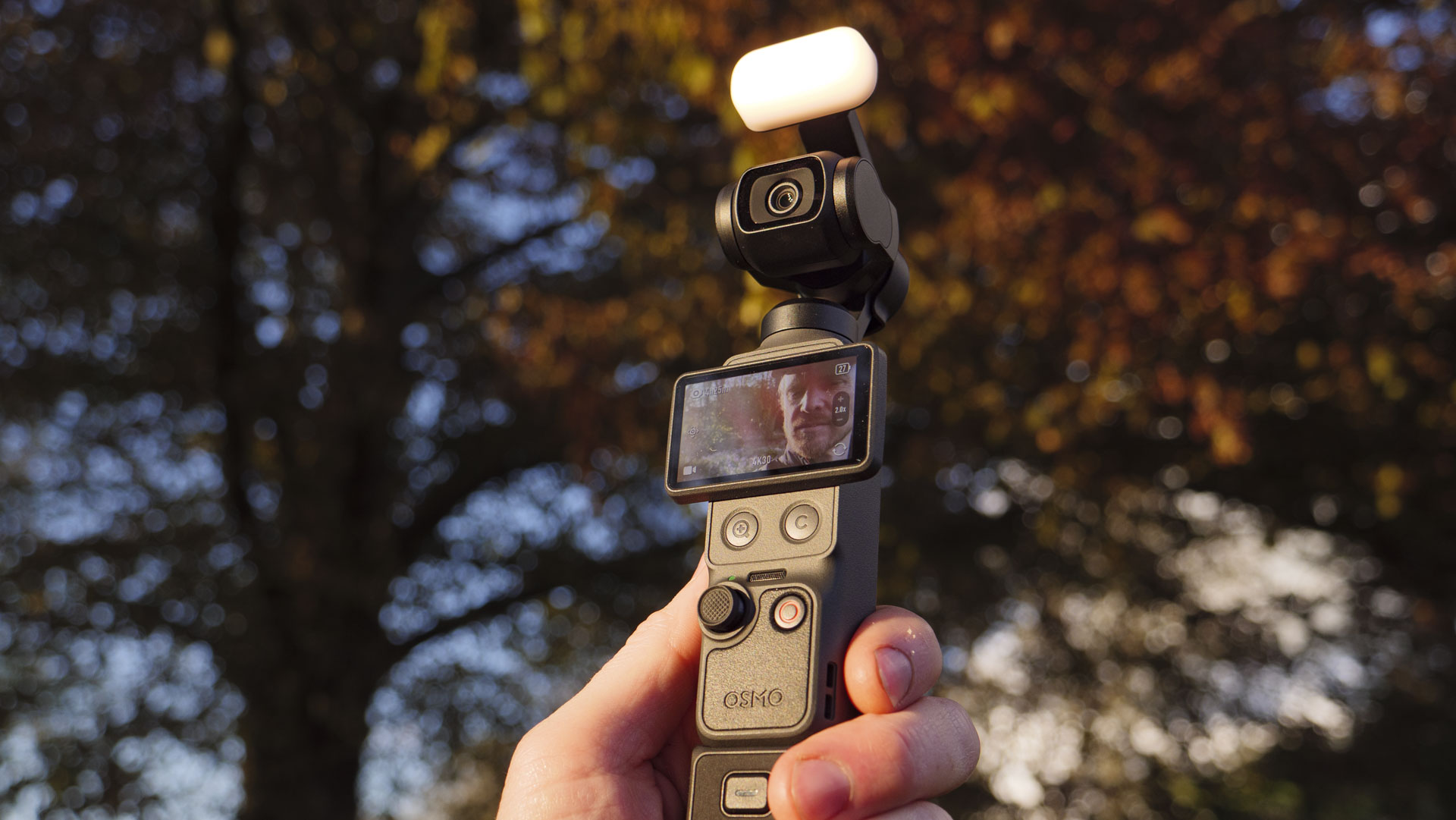 DJI Osmo Pocket 4 vlogging camera with natural background at golden hour, and the user in the frame of the screen