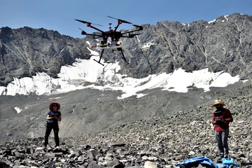 Two people standing on a rocky surface with mountains in the background. The person on the right is holding a controller, controlling the drone in the middle of them above the ground.