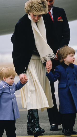 British Royal Diana, Princess of Wales (1961-1997), wearing a grey, black and white sweater under a black jacket with a cream scarf and cream skirt, holding the hands of her sons, Prince Harry and Prince William, after their arrival at Aberdeen Airport in Aberdeen, Aberdeenshire, Scotland, 14th March 1986