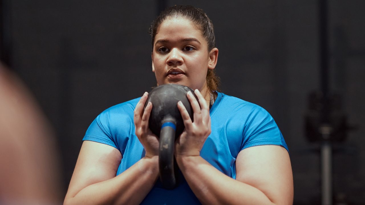 Woman in blue T-shirt holds kettlebell by her chest in two hands