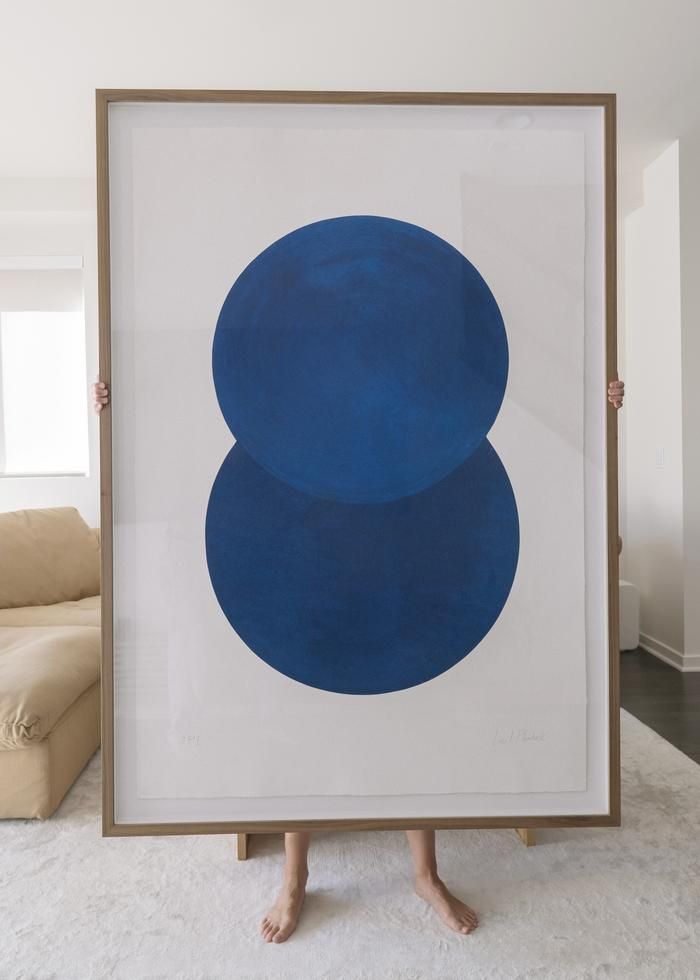 A person holding a larger-than-life frame containing a white and blue abstract print in a palely decorated living room.