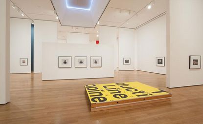 Christopher Williams's exhibition at MoMa in New York. Photographs are hung low and spaced out. On the floor, there is an art piece painted yellow with writing on it.