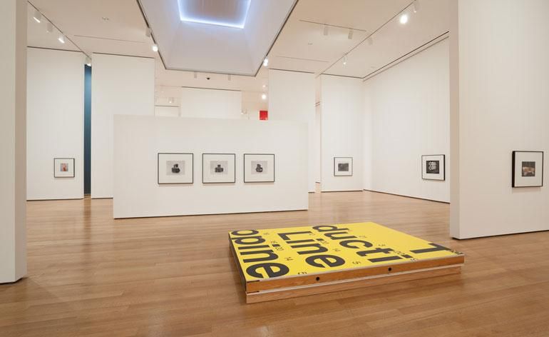 Christopher Williams&#039;s exhibition at MoMa in New York. Photographs are hung low and spaced out. On the floor, there is an art piece painted yellow with writing on it.