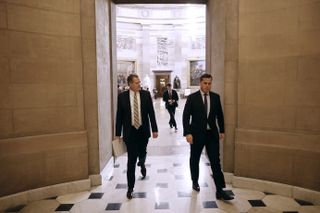 two men in suits walk through a hallway in a building filled with sculptures and paintings