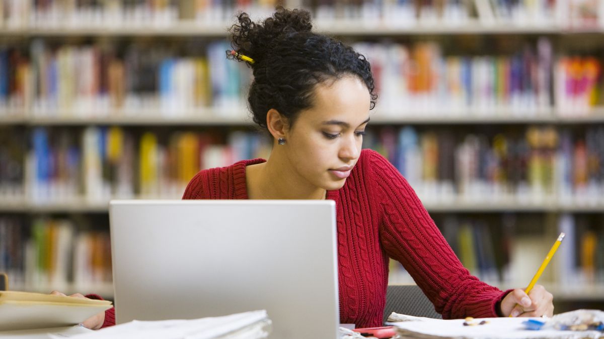 Female computing student working at a desk in a library with laptop.