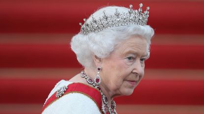 Queen Elizabeth wearing a white dress, red sash, diamond and ruby jewelry and a diamond tiara in front of a red staircase