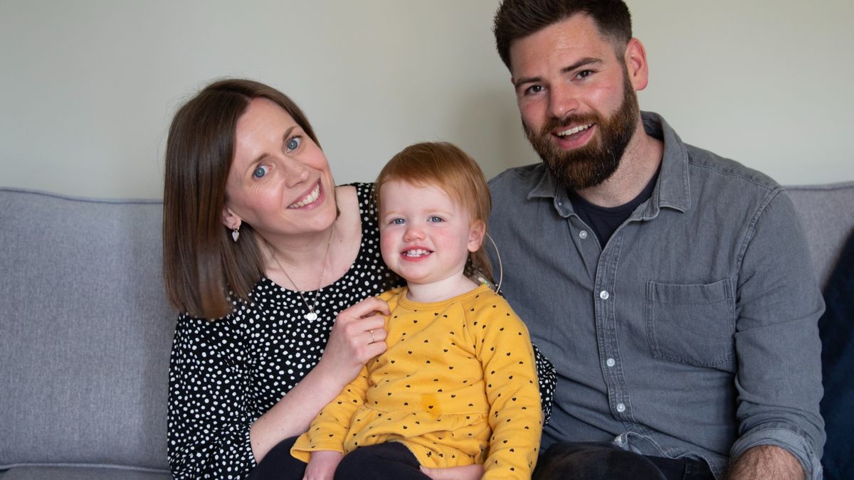 Image of a toddler girl sat with her mother on her left and her father on her right. They are all smiling at the camera. The mother is wearing a black-and-white polka dot top, the toddler is wearing a bright yellow top and the father is wearing a grey shirt.