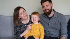 Image of a toddler girl sat with her mother on her left and her father on her right. They are all smiling at the camera. The mother is wearing a black-and-white polka dot top, the toddler is wearing a bright yellow top and the father is wearing a grey shirt.