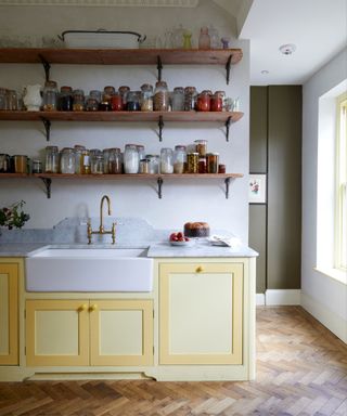 Kitchen with white walls, parquet flooring, yellow cupboards with inset butler's sink, reclaimed wooden shelving above with glass storage jars.