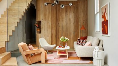 A living room with a wood staircase, wood walls, a vase of flowers, a white side table, a colorful rug, a floor lamp, framed paintings, and a gray couch
