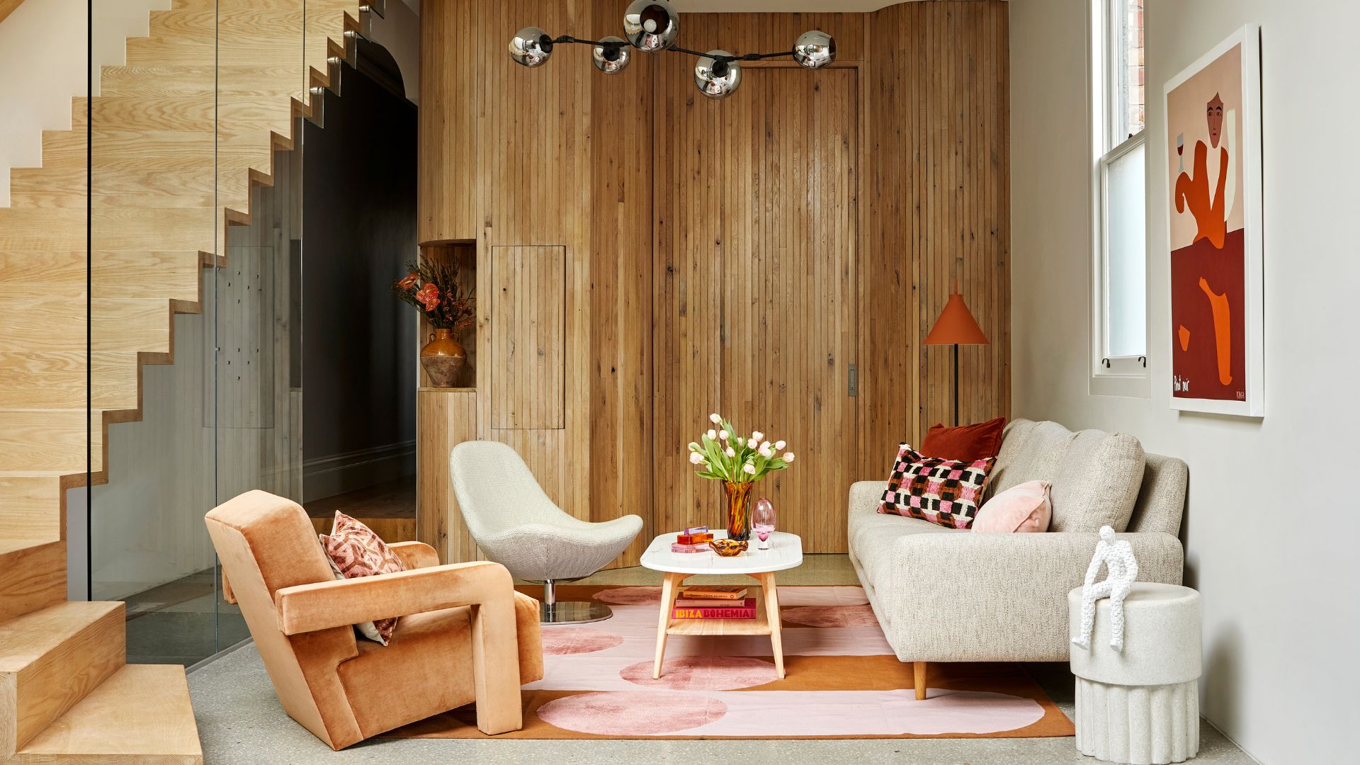 A living room with a wood staircase, wood walls, a vase of flowers, a white side table, a colorful rug, a floor lamp, framed paintings, and a gray couch