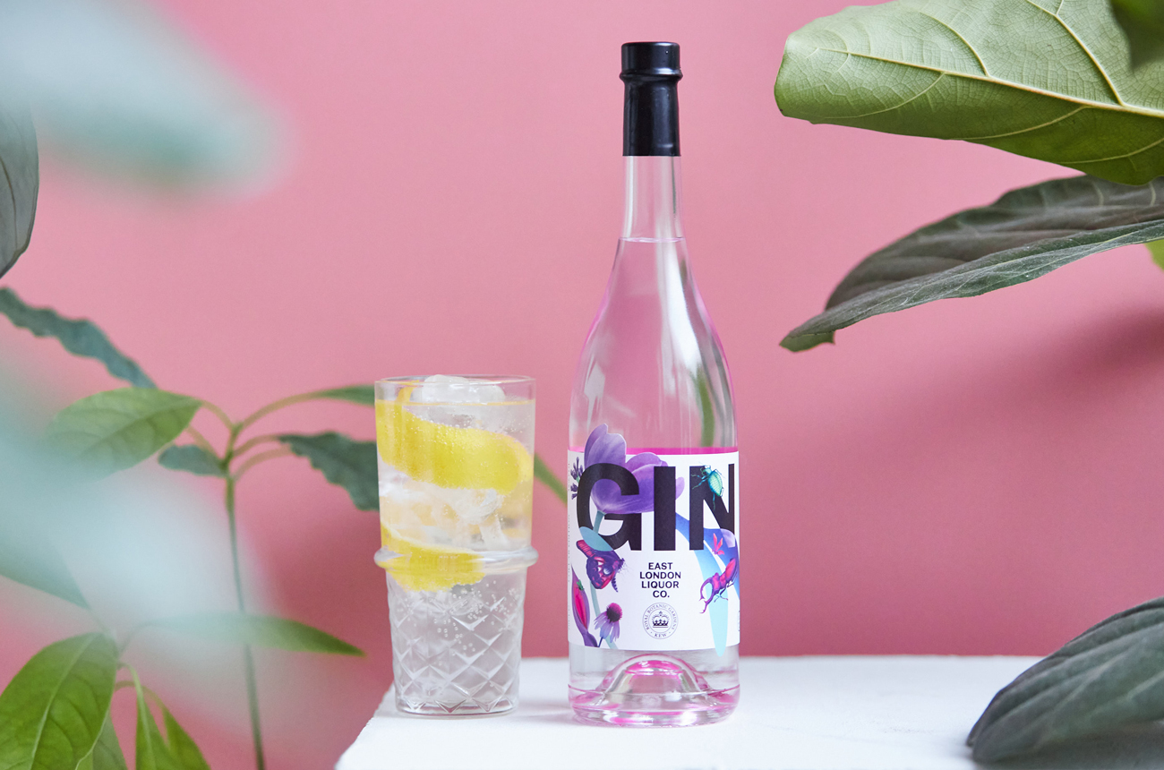 A bottle of gin and glass of gin and tonic on a white table with a pink background and leaves